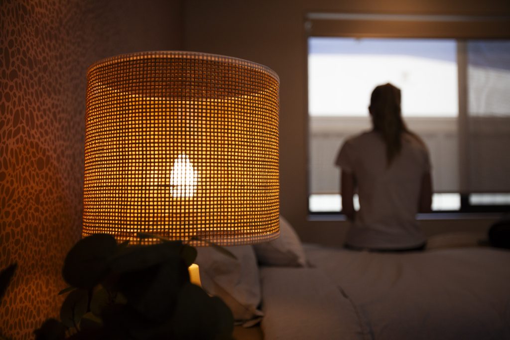 Image of woman sitting on the edge of a bed looking out of a window and a bedside lamp in the forefront.