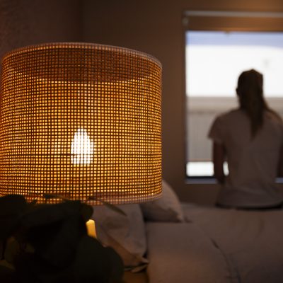 Image of woman sitting on the edge of a bed looking out of a window and a bedside lamp in the forefront.