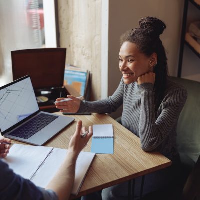 Cheerful young stylish woman at meeting with male partner in cafe. Discussing startup at laptop.